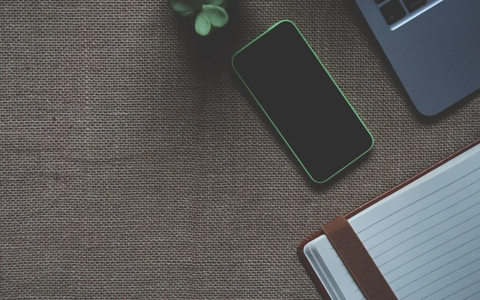 A smartphone, a notebook and a small plant on a burlap desk, seen from above