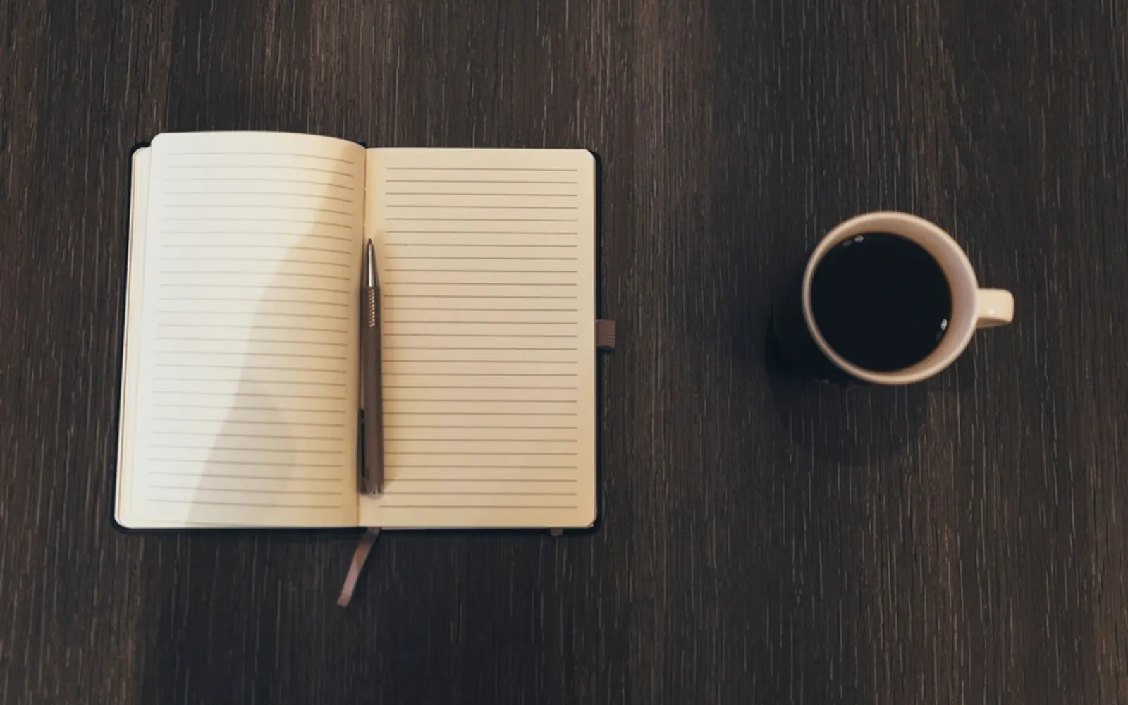 An open notebook and pen next to a cup of coffee on a dark wooden table