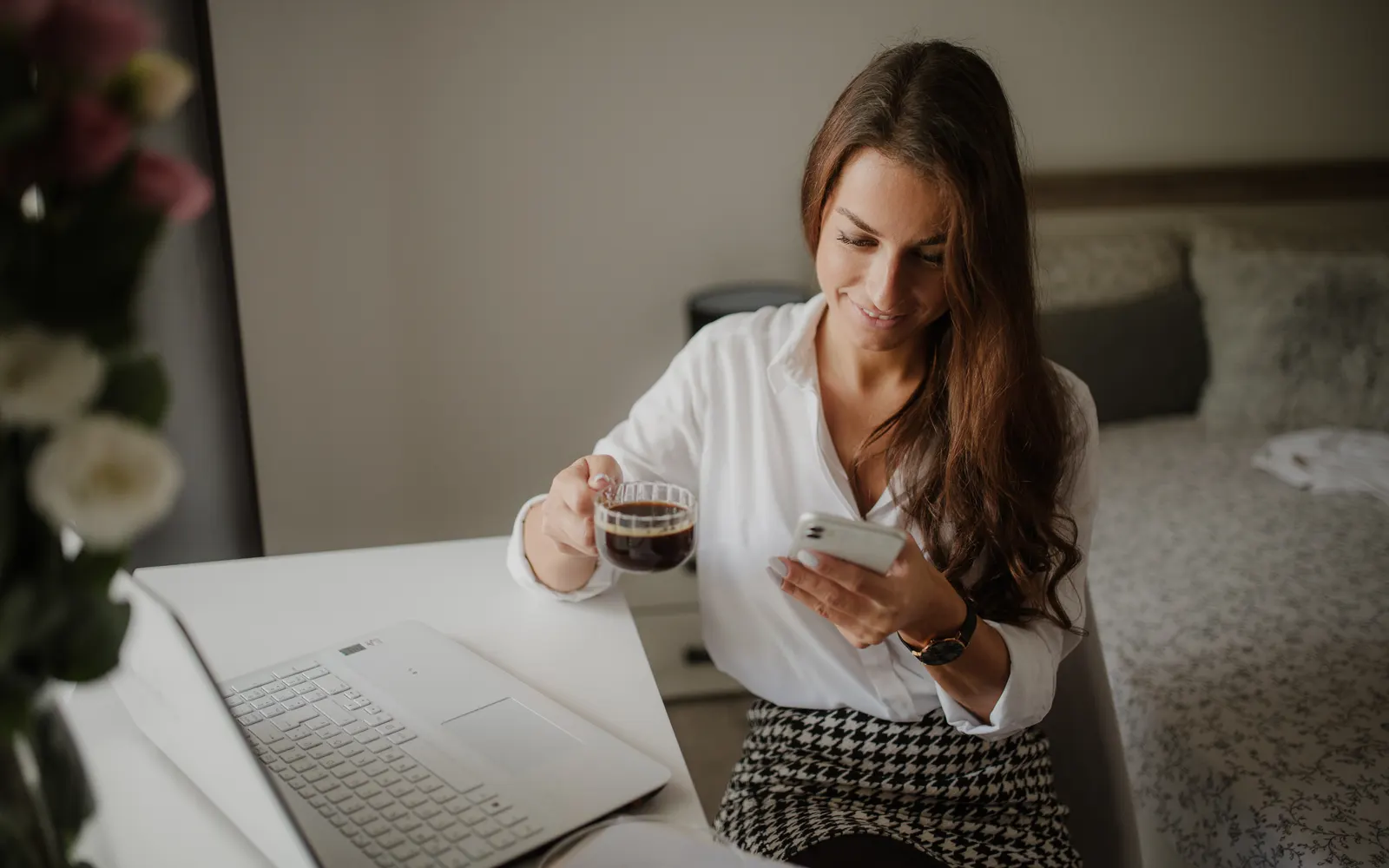 A woman in a white shirt holding a glass of coffee and smiling at her phone at her desk