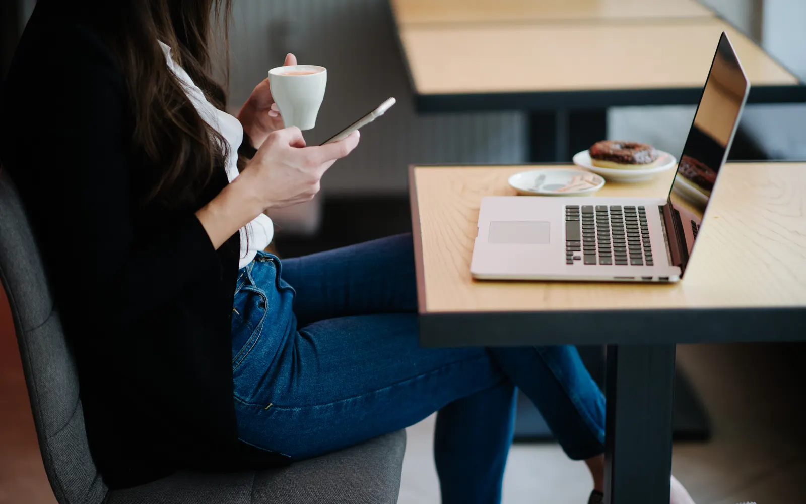 A woman in a cafe holding a coffee cup and her phone, with a laptop on the table beside her