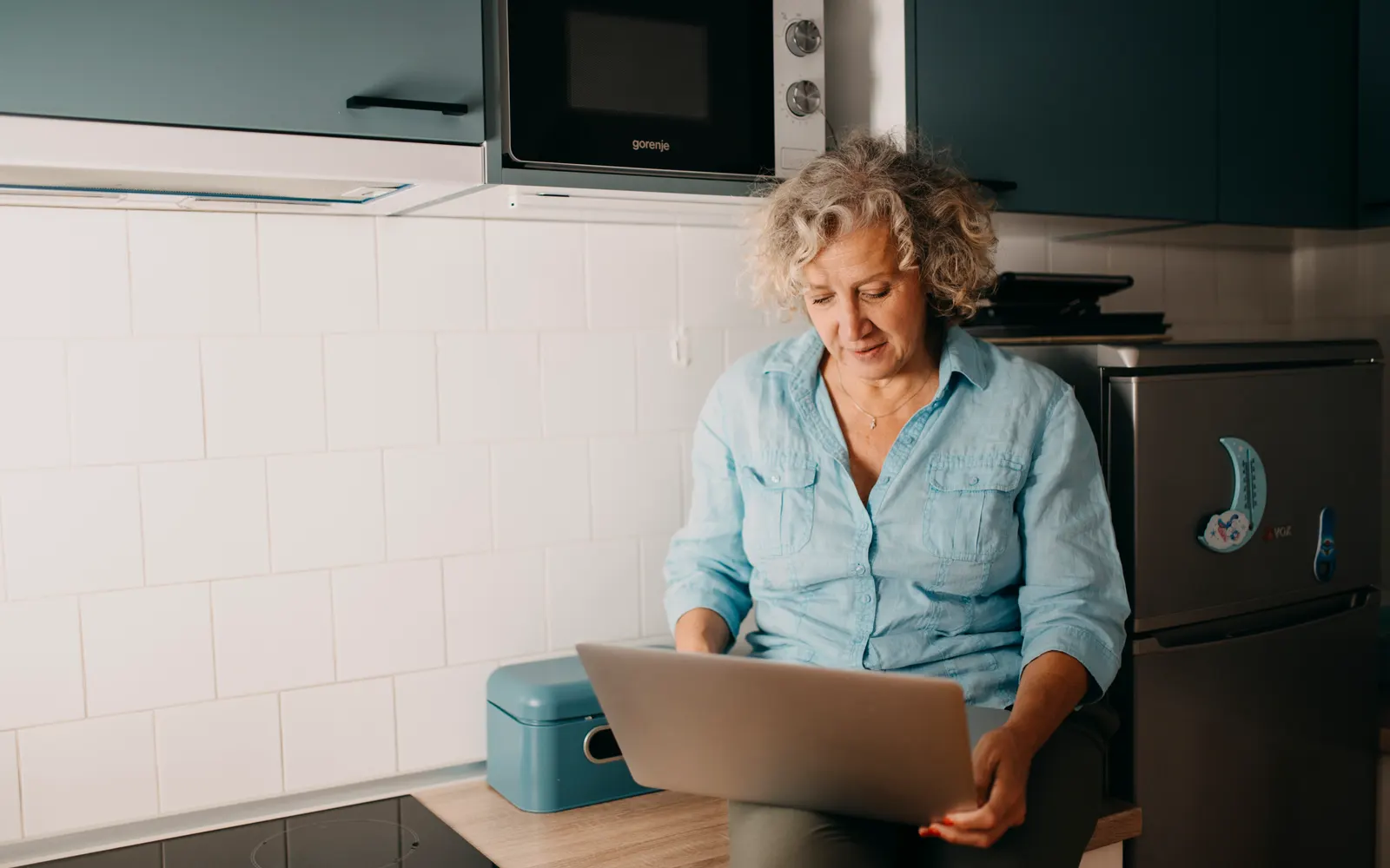 A woman with grey curly hair working on a laptop in her kitchen