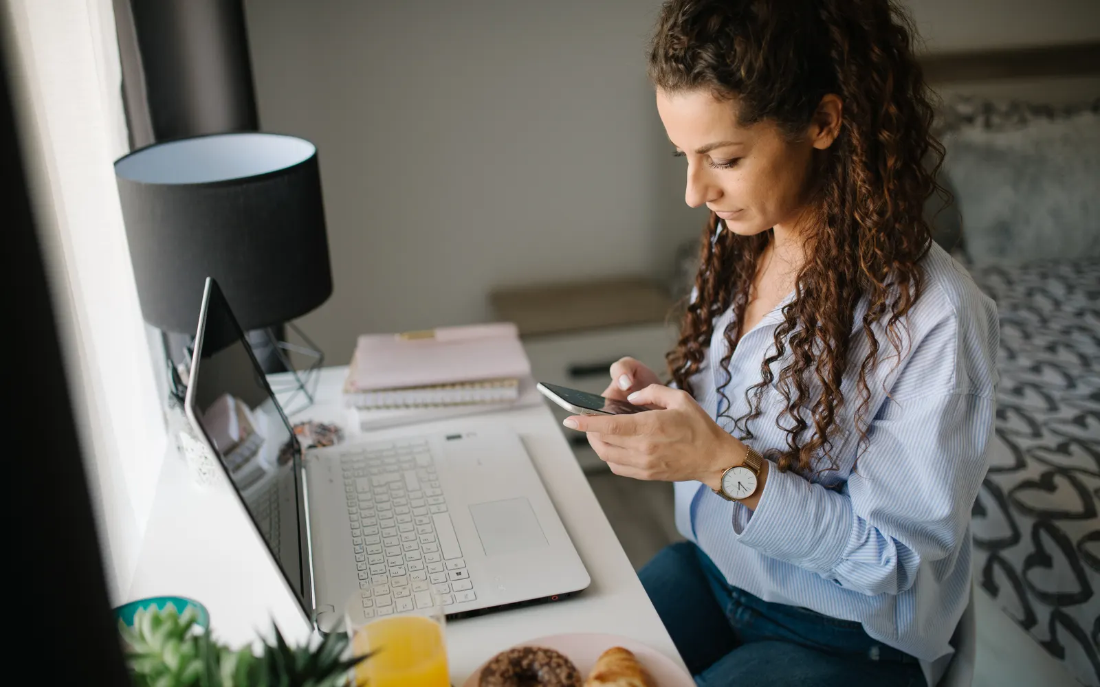 A woman with curly hair checking her phone at a desk beside her laptop